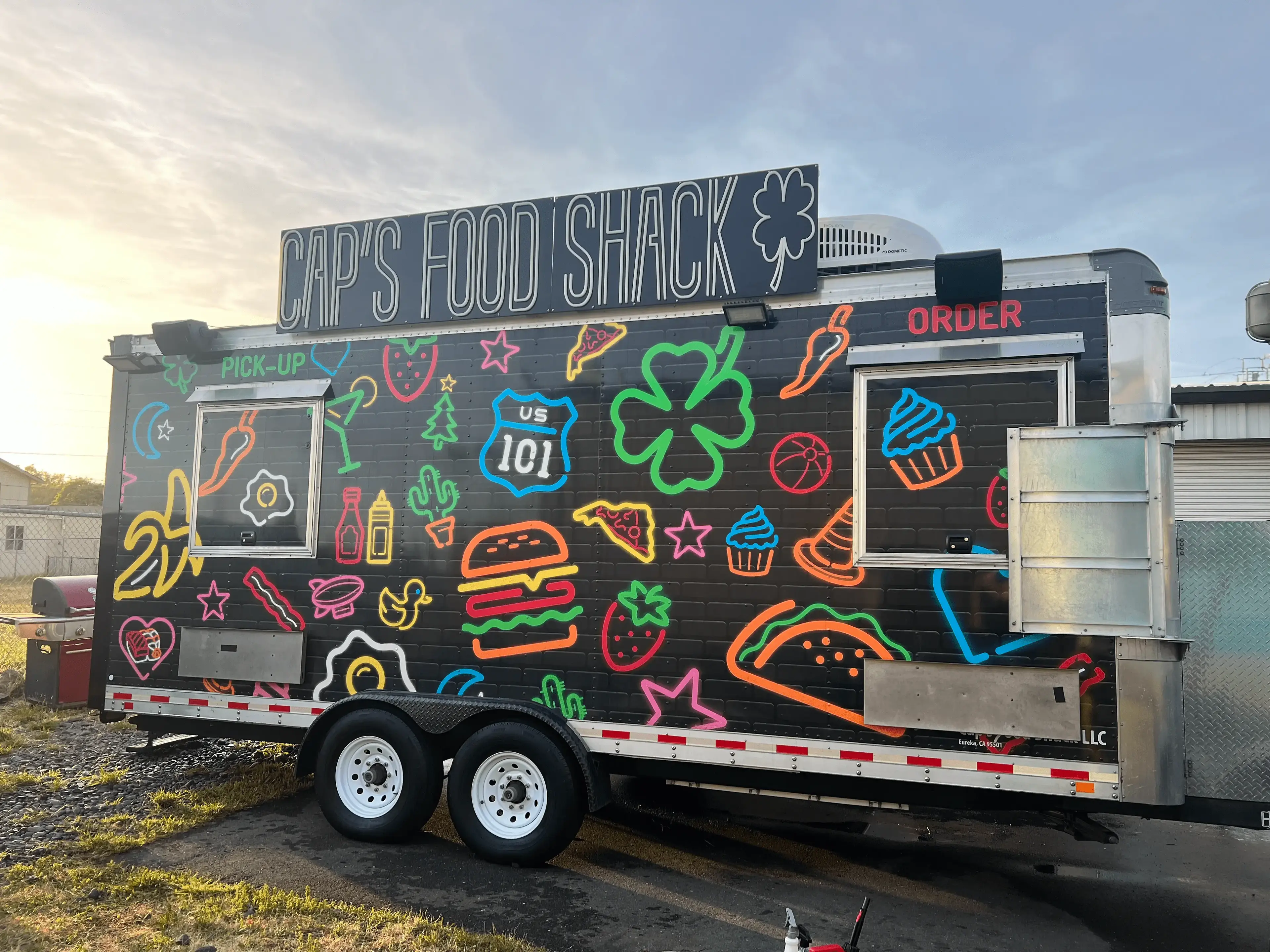 Cap's Food Shack food truck with colorful neon graphics of food items and signs, set against a sunset sky, ready for pick-up and order service.